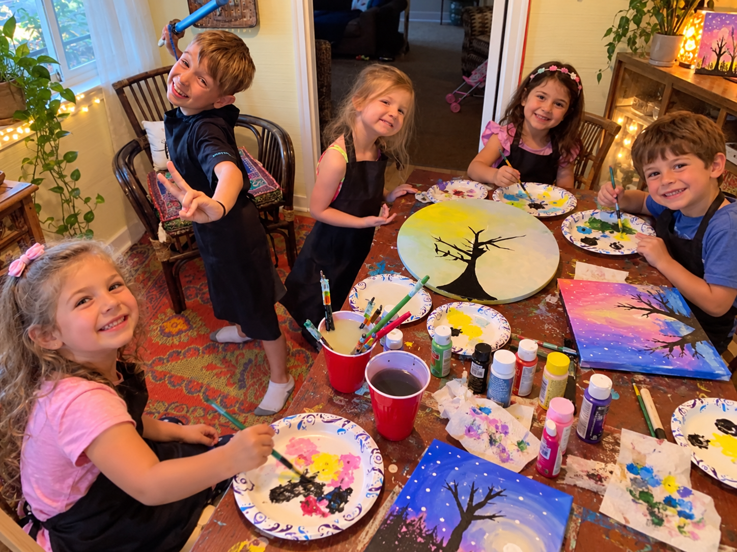 Five smiling children gathered around a craft table covered with painted canvases, paint bottles and brushes