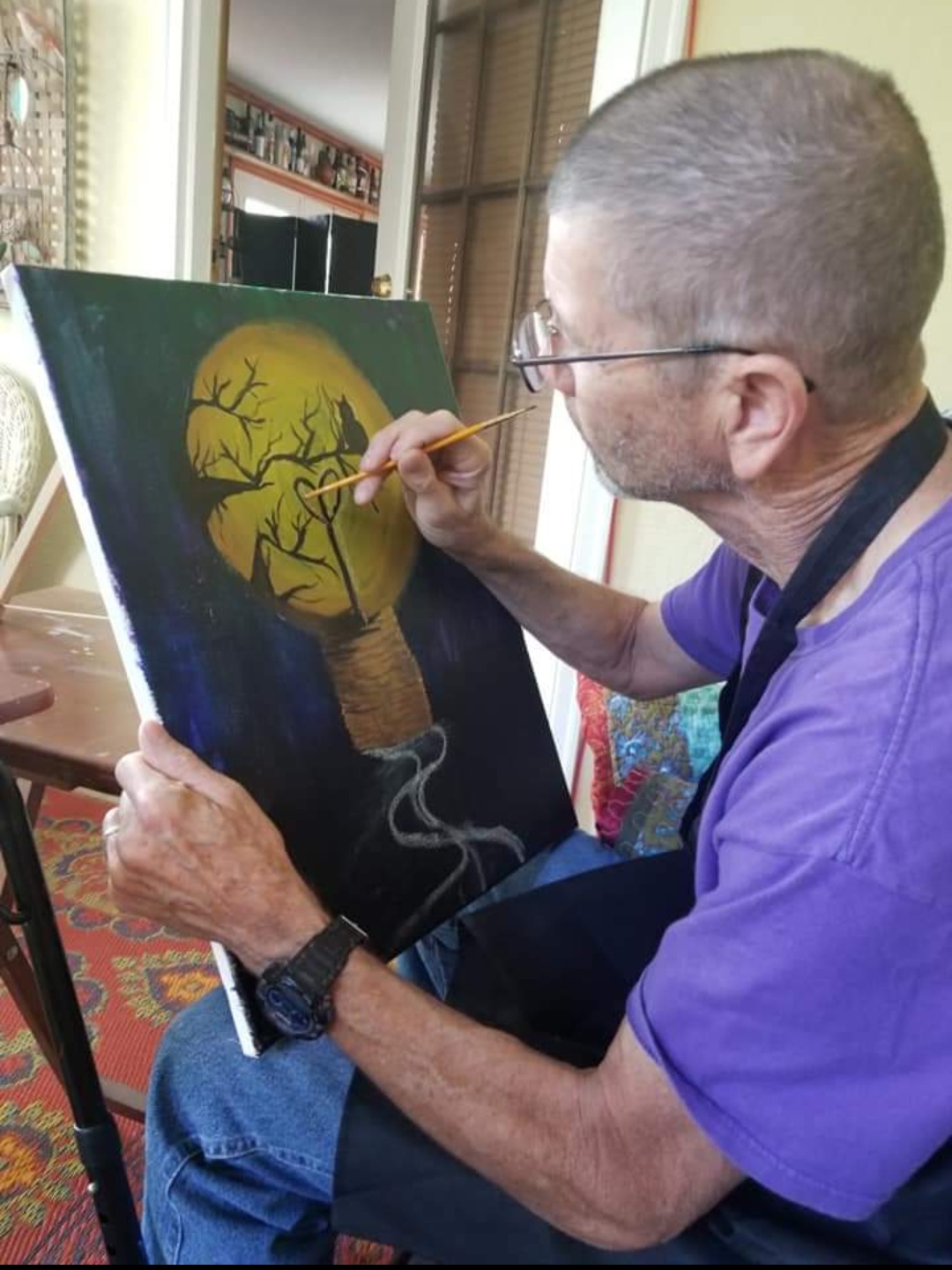 An adult man in an apron painting a glowing moon scene on a canvas