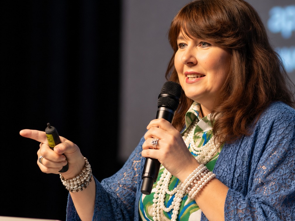Michelle speaking confidently on stage, holding a microphone and gesturing with a clicker in her other hand