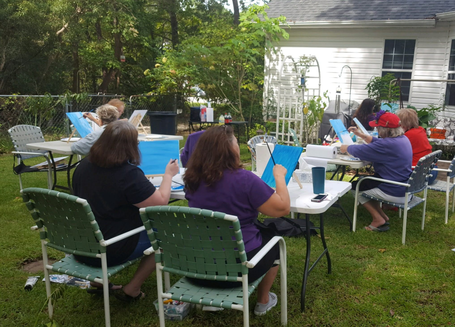 Outdoor painting class on a lawn, adults seated at white tables painting on tabletop easels